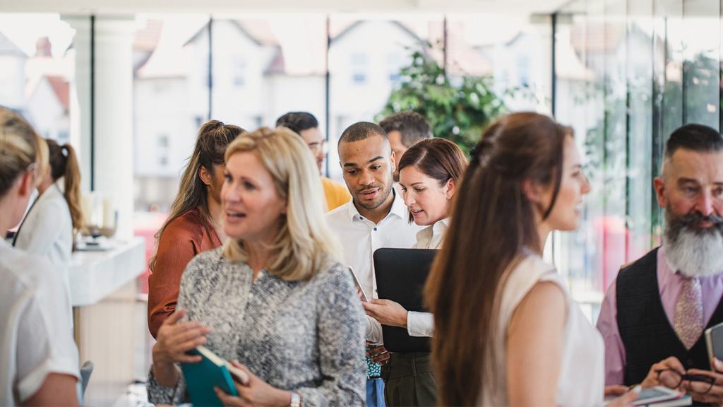 A bustling networking event with a diverse group of people engaging in conversations. Some individuals hold notebooks, while others use laptops or tablets. The background features large windows allowing natural light to flood the modern, airy space.