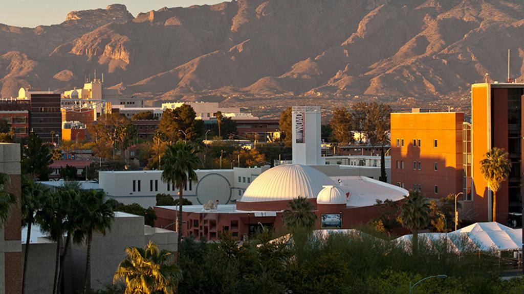 A cityscape showing the University of Arizona campus buildings with the white domed observatory in the center. In the background, the sunlit Catalina Mountains rise prominently against a clear sky. Palm trees and other greenery are interspersed throughout the campus.