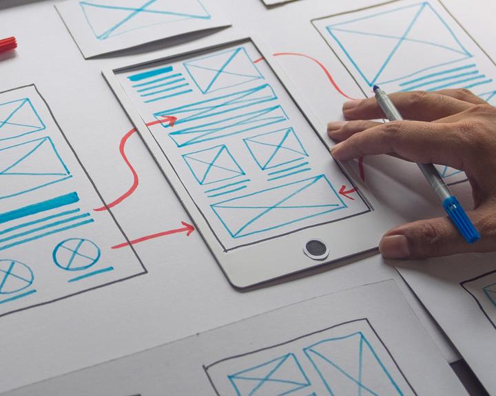 A hand holding a pen sketches a mobile app prototype on paper. Several papers with wireframes, including screens with text blocks, images, and buttons, are spread out. Red arrows indicate navigation between different screens.