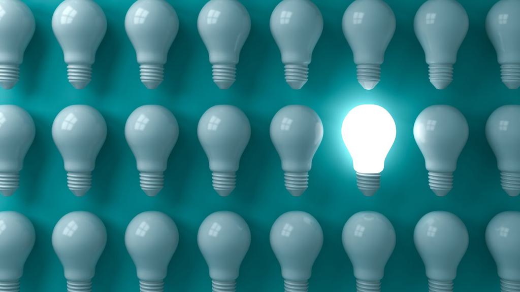 A grid of twenty-four unlit light bulbs against a teal background, with one bulb in the middle of the image brightly lit. The glowing bulb stands out in sharp contrast to the others, emphasizing its uniqueness.