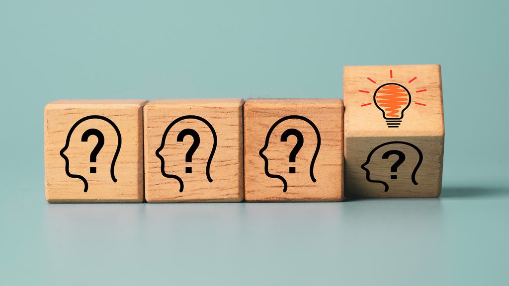 Four wooden blocks are lined up against a teal background. The first three blocks show a profile of a head with a question mark inside. The fourth block shows a lit-up light bulb inside a head, signifying the idea or solution.