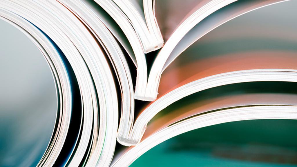Close-up photograph of several open magazines stacked on top of each other, with pages slightly curved and creating abstract overlapping shapes. The image focuses on the edges and curves of the pages, showcasing a colorful and artistic arrangement.