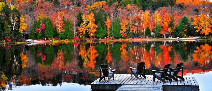 A serene lakeside scene with brightly colored autumn trees reflected in the calm water. Three wooden chairs are set on a wooden deck extending over the lake, offering a peaceful spot to enjoy the vibrant foliage. A few scattered houses are visible among the trees.