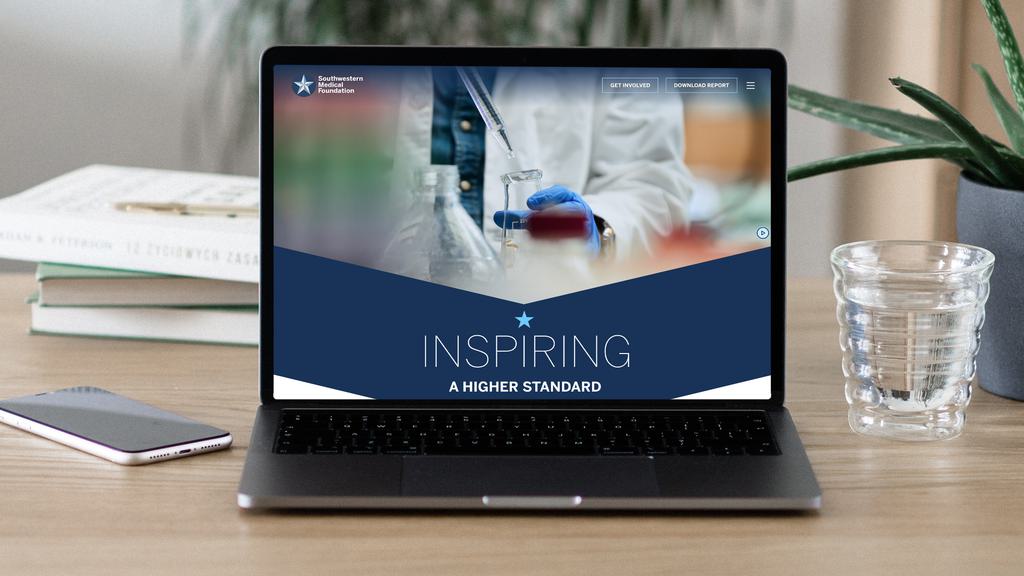 A laptop on a desk displays a website with the heading "Inspiring A Higher Standard" and an image of a scientist working in a lab. The desk also has a smartphone, a clear glass of water, and a small stack of books.
